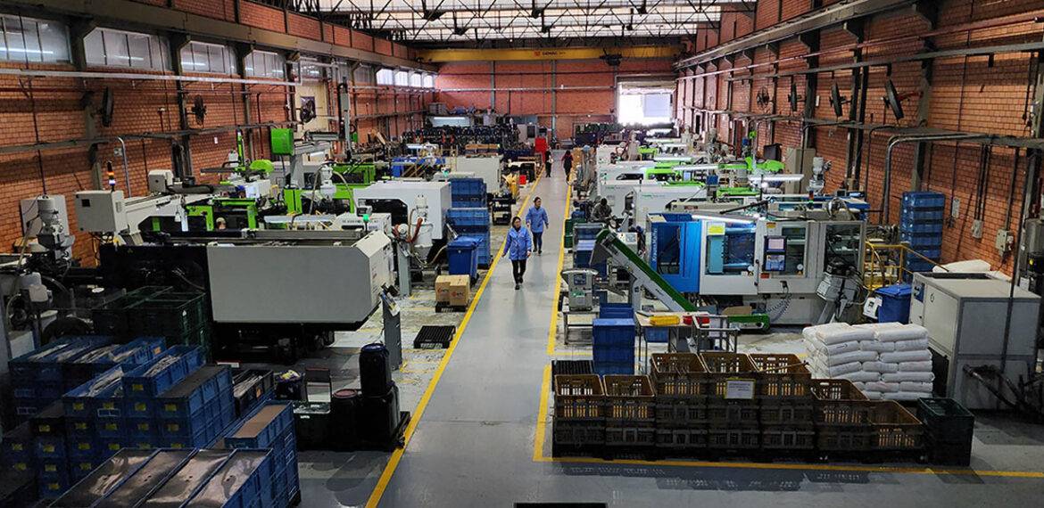 Vista ampla do setor de injeção plástica da NTC Company, com diversas máquinas injetoras em operação, operadores circulando pelo corredor central e organização de insumos e peças ao redor das células produtivas.