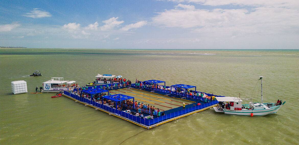 Vista aérea da piscina flutuante do evento AquaRace 2025, instalada sobre uma plataforma modular Pierplas no mar de João Pessoa (PB). A estrutura retangular, cercada por áreas azuis e repleta de espectadores, abriga atletas e equipes durante as provas de natação, com barcos de apoio ao redor.