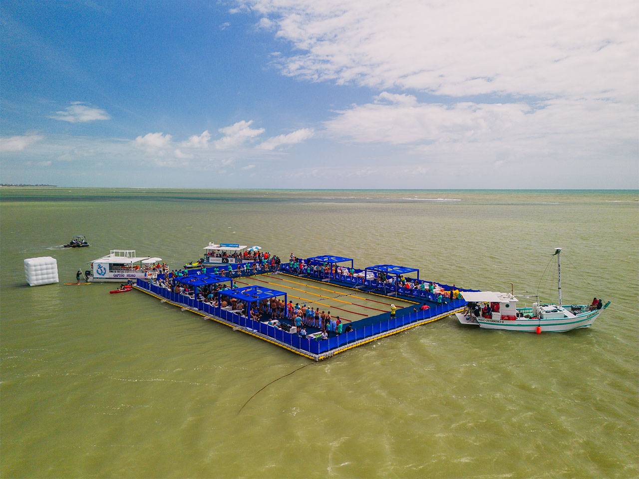 Vista aérea da piscina flutuante do evento AquaRace 2025, instalada sobre uma plataforma modular Pierplas no mar de João Pessoa (PB). A estrutura retangular, cercada por áreas azuis e repleta de espectadores, abriga atletas e equipes durante as provas de natação, com barcos de apoio ao redor.