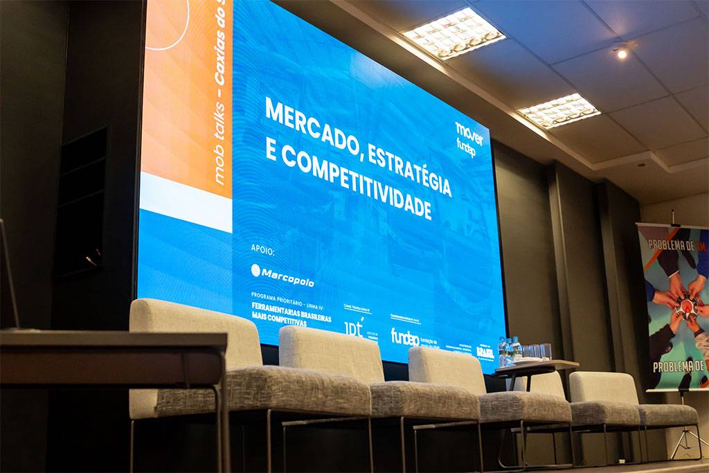 Painel de auditório preparado para o evento Mob Talks sobre “Mercado, Estratégia e Competitividade”, com cadeiras posicionadas no palco diante de uma grande tela. Não há pessoas no palco.