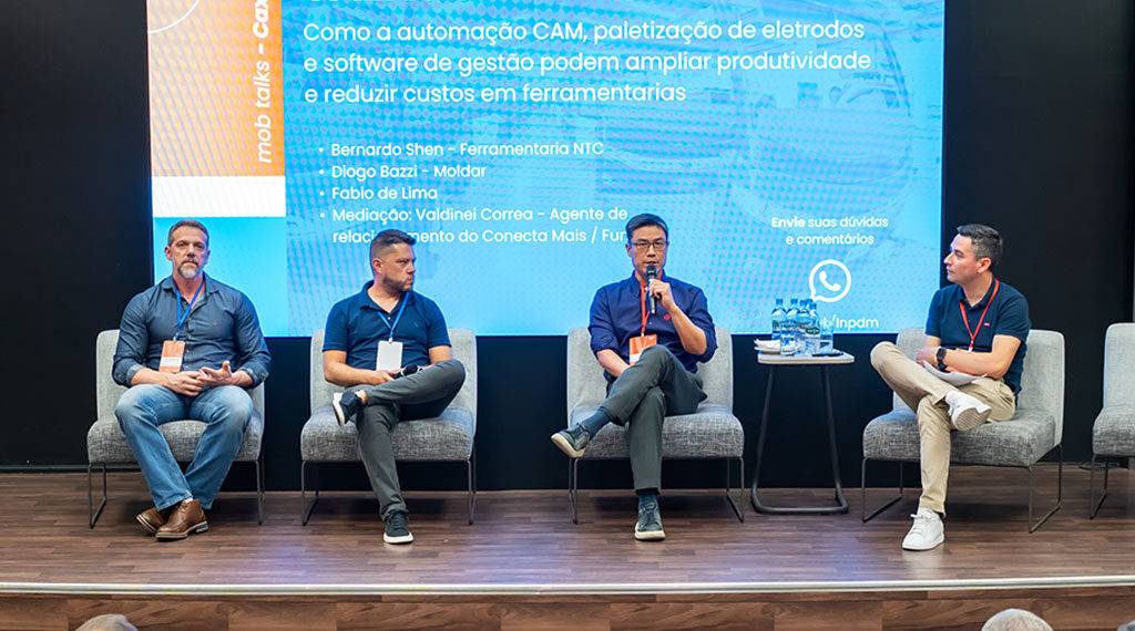 Plateia acompanhando o painel com quatro palestrantes no palco. O diretor Bernardo Shen, da NTC, está na terceira posição da esquerda para a direita, participando da discussão. A grande tela ao fundo exibe o conteúdo do evento.