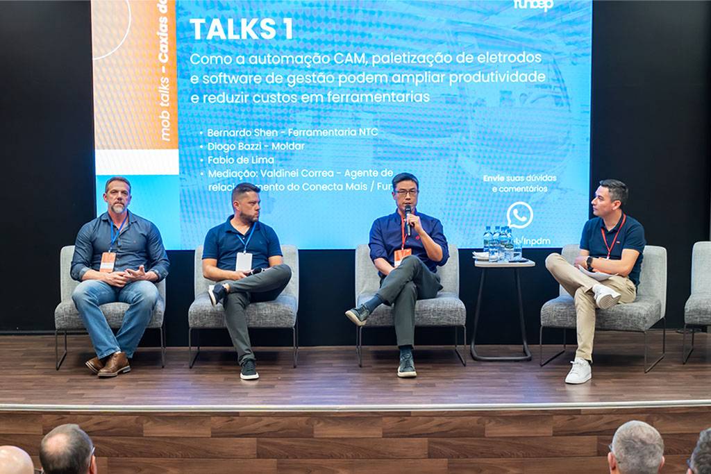 Plateia acompanhando o painel com quatro palestrantes no palco. O diretor Bernardo Shen, da NTC, está na terceira posição da esquerda para a direita, participando da discussão. A grande tela ao fundo exibe o conteúdo do evento.