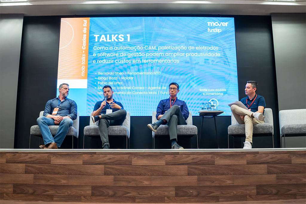 Imagem centralizada do painel com quatro participantes no palco. O diretor Bernardo Shen, da NTC, está na terceira posição da esquerda para a direita, sentado e atento à conversa. A tela exibe o título “Talks 1” e o tema técnico do debate.