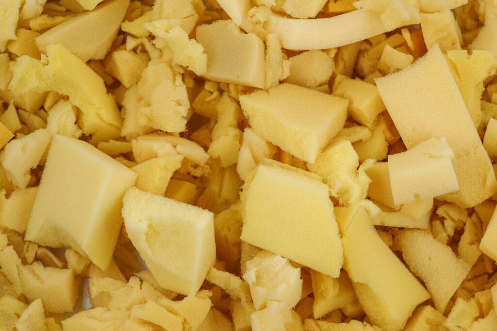 Uma vista superior de diversos pedaços irregulares e retalhos de espuma de poliuretano flexível em tons de amarelo claro e bege. Os pedaços variam em tamanho e formato, amontoados de forma aleatória, evidenciando a textura macia e esponjosa do material frequentemente usado em estofados e colchões.