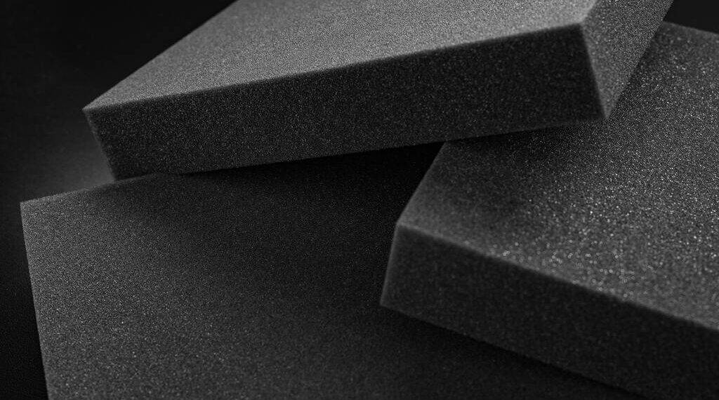 Fotografia em close-up de três blocos retangulares de espuma de poliuretano expandido na cor cinza escuro (grafite). Os blocos estão sobrepostos de forma assimétrica contra um fundo preto. A iluminação lateral destaca a textura porosa e granular da superfície da espuma.